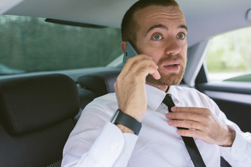 Indoor portrait of a handsome businessman calling on smart phone straightens black tie and sitting on car back seat. Technology and business concept.