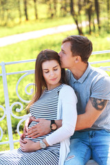 Handsome man and his beautiful pregnant wife are hugging and smiling while standing in park