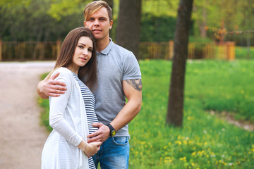 Handsome man and his beautiful pregnant wife are hugging and smiling while standing in park