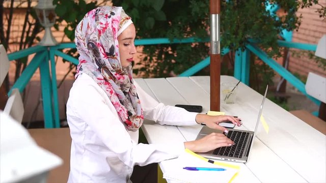 A modern and young Muslim woman in a hijab on her head prints a message for her friends on a laptop, a lady in a scarf with a smile writes text on the keyboard