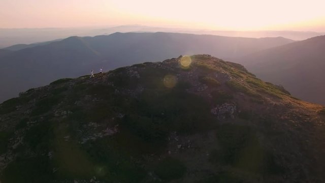 Drone footage of young adventurous couple standing on top of mountain after long hike, overlooking beautiful purple sunset, hugging and kissing with hair blowing in wind on rock cliff high altitude