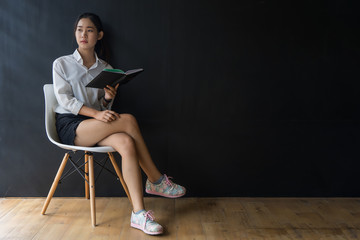 Asian woman thinking with a book and sitting on a chair.