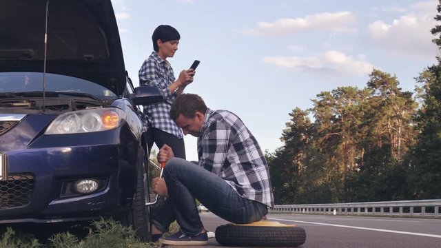 handsome caucasian man changing a tire on the side of the road. Woman using mobile phone messaging or scrolling social media on smartphone .