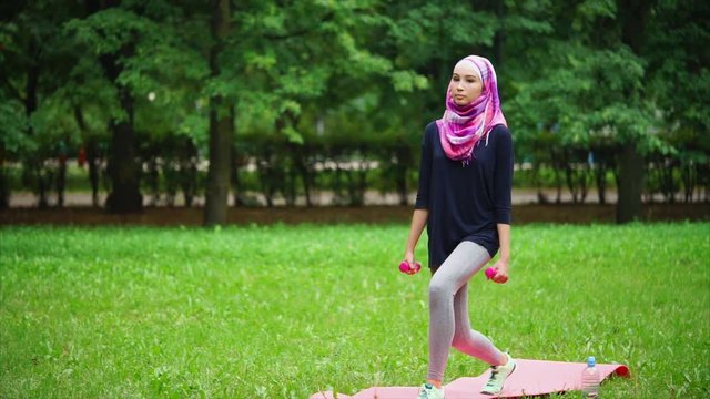 Young muslim woman doing exercises in the park summer day, a lady in a shawl squats with dumbbells in her hands, she supports her body in good physical shape