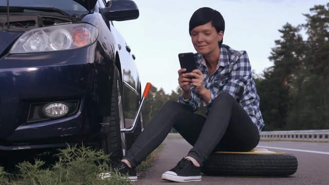 Woman stuck on the side of the road with a flat tire. Caucasian driver sitting on tire at the roadside holding smartphone messaging with friend of surfing internet.
