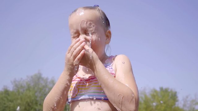 Cute baby girl running through the fountain in a public Park and laughs. water leaked in the eyes and she frown and spits