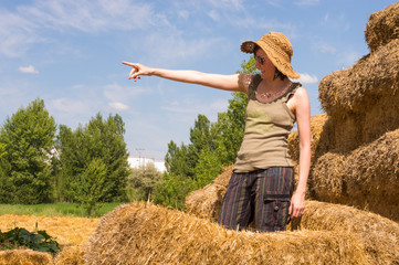 Naklejka na ściany i meble Pretty woman with hat standing in straw bales and pointing her finger toward the blue sky.