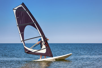 Windsurfer man in wetsuit windsurfing on board with colorful sail in calm sea or ocean on sunny day on blue sky background. Recreational water sports during idyllic summer vacation