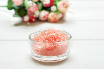Pink sea salt in glass bowl with rose flowers behind on a white wooden table