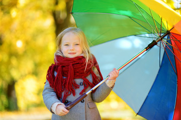 Cute little girl holding rainbow umbrella on beautiful autumn day. Happy child playing in autumn park.