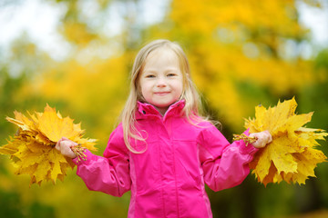 Cute little girl having fun on beautiful autumn day. Happy child playing in autumn park. Kid gathering yellow fall foliage.