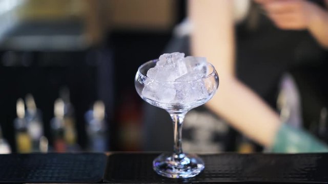 Close up of an unrecognizable woman bartender putting ice cubes into a cocktail glass and placing it on the counter. Handheld real time close up shot
