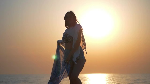 Carefree woman dancing in the sunrise on the sea beach. Vacation vitality healthy living concept. Girl Holding Scarf and Spinning around. Femininity At Sunset, Slow motion.