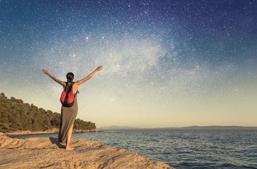 Fototapeta na wymiar A young woman on the seashore raises her hands to the sky against the starry sky.