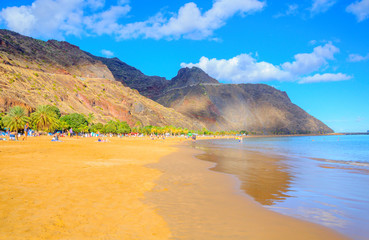 beach on the island of Tenerife in bright sunny day, a great number of vacationers ashore, on a background mountains tower