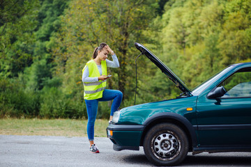 Worried young white female holding her head staring at the car engine of her broken car waiting for repair service