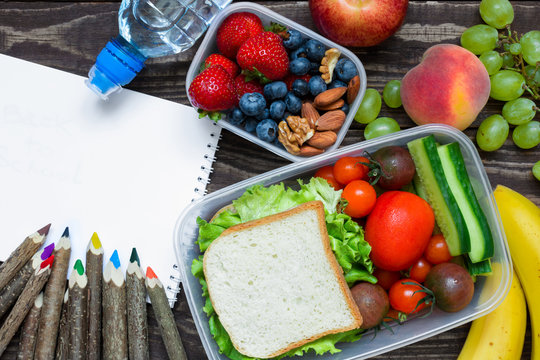 school lunch boxes with sandwich, fruits, vegetables and bottle of water with colored pencils and empty copybook