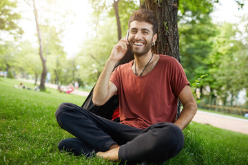 Naklejka na ściany i meble Cheerfully smiling to the camera bearded guy in red t-shirt sitting cross-legged under tree. Caucasian man with stylish haircut talking on smartphone. Positive emotions and communication.