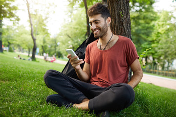 Naklejka na ściany i meble Attractive good-looking caucasian man looking with smile on the screen of his mobile phone, happy because of chatting with his girlfriend. Hipster casually weared sitting cross-legged on the grass