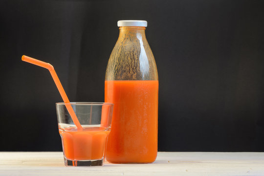 A bottle of carrot juice and a glass of carrot juice with a straw on white wood table on black background. Still life of a healthy eating concept