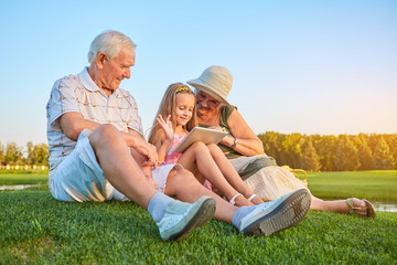 Smiling girl with grandparents, tablet. Happy people sitting on grass. How internet makes life easier.