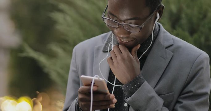 Young african student in classical suit listening to music with earphones outdoor