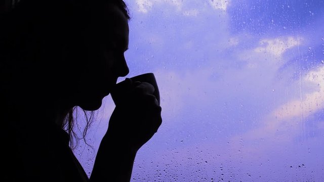 Silhouette of a girl with a cup by the rainy window. Girl with a cup of coffee by the window.