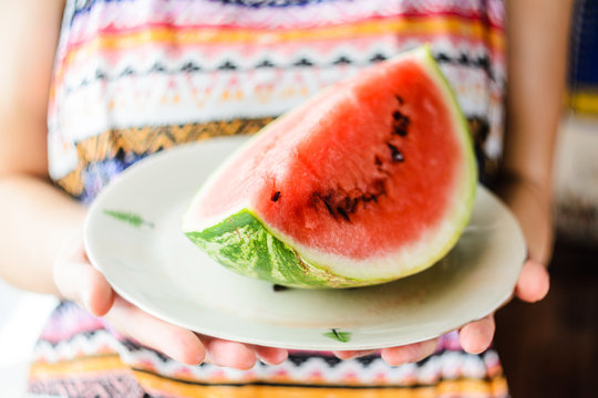 Watermelon background. Girl in a colorful summer shirt holding a plate with a large slice of watermelon in the sun