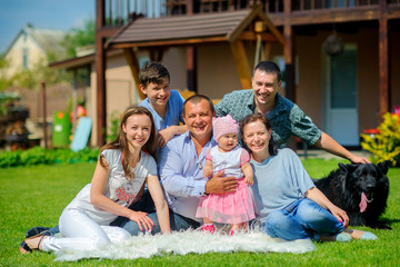 A large, friendly family sits on the grass near their house, hugs and smiles tightly, next to them sits a dog