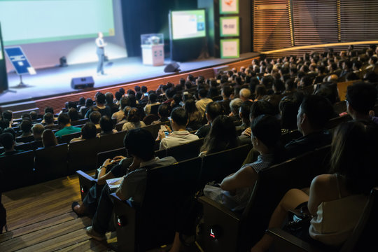 Rear View Of Audience Listening The Speakers On The Stage In The Conference Hall Or Seminar Meeting With Low Light Situation, Business And Education About Investment Concept