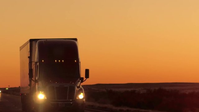 CLOSE UP LENS FLARE: Semi truck driving and hauling goods on empty highway across the Great Plains in golden morning. Freight delivery truck transporting cargo on interstate freeway at stunning sunset