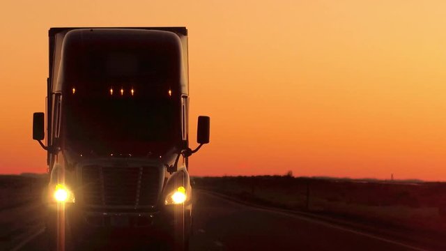 CLOSE UP LENS FLARE: Semi truck driving and hauling goods on empty highway across the Great Plains in golden morning. Freight delivery truck transporting cargo on interstate freeway at stunning sunset