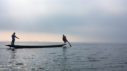 Black silhouette of two fishers standing on stern and paddling by legs, travelling by traditional long boat on Inle lake, Myanmar. Culture and traditions of Burmese people. Asian travel background.