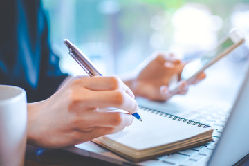 Business women hand are working at a notebook computer and taking notes in a pen notepad in the office.Soft focus