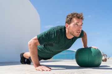 Pushup bodybuilder athlete strength training chest and shoulder muscles doing single arm alternating medicine ball push-ups floor exercises at outdoor gym.