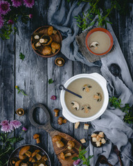 mushroom soup in white bowl with mushroom slices with wooden board, raw mushrooms, green leaves and flowers, gray textile, wooden spoons, dry bread slices on gray wooden background, top view