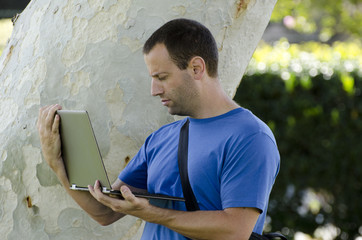 Man working on his lap top outside in front of a tree trunk.