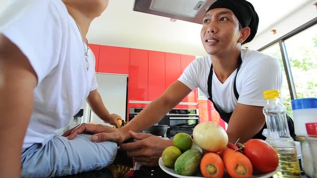 Private Thai chef cooking, his little son in chef's hat nearby sitting on the table in a modern style home kitchen. Making Thai food: mixed vegetables oyster sauce