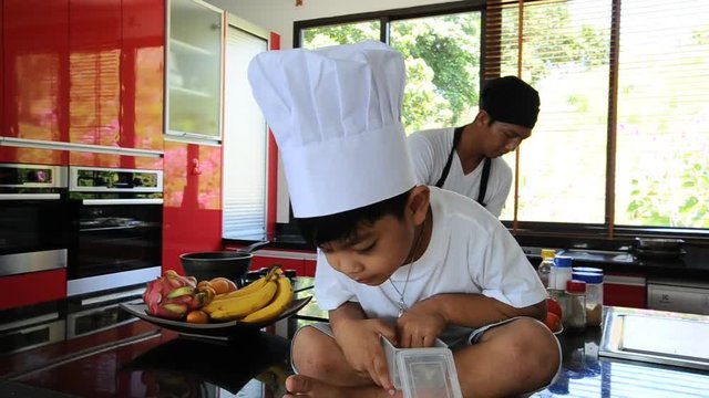 Private Thai chef cooking, his little son in chef's hat nearby sitting on the table in a modern style home kitchen. Making Thai food: mixed vegetables oyster sauce