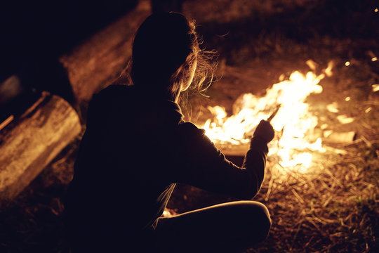 Young woman in geans and hoodie in the mountains. Young woman with fire on mountain at sunset. Girl standing near a fireplace, back view