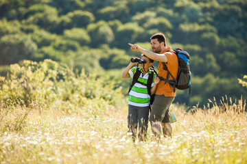 Father and son are hiking in nature in summer. They are using binoculars. 