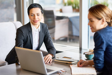 Multi-ethnic group of coworkers wearing suits gathered together at cozy cafe and brainstorming on joint project, panoramic window on background