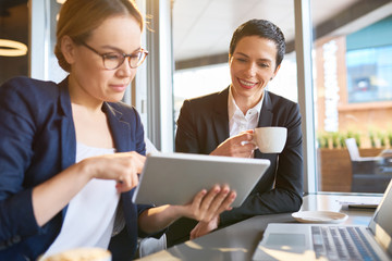 Pretty Asian manager in eyeglasses showing results of accomplished work to her smiling superior with help of digital tablet while having working meeting at cozy coffeehouse.