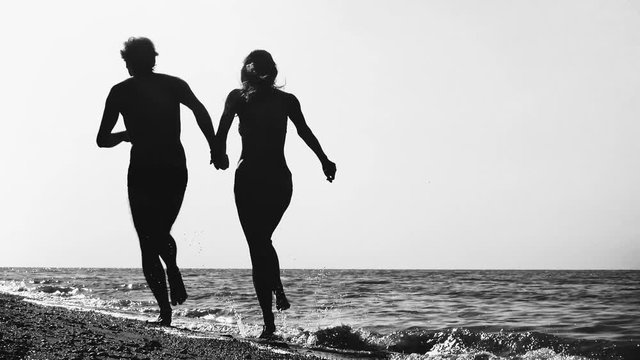 Back view of young man-woman couple in love enjoys and run along ocean beach sea-side at sunrise or sunset in summer. black and white slow motion