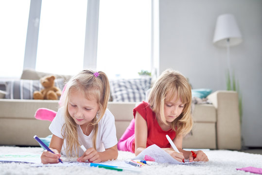 Portrait of two little girls drawing and coloring pictures lying on floor in cozy living room