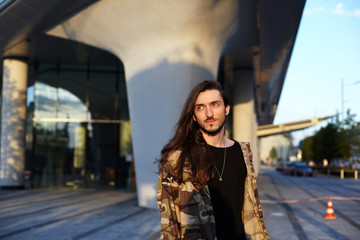 Urban fashion portrait of attractive trendy-looking young bearded Caucasian man dressed in camouflage bomber over black t-shirt, having nice walk on city streets with friends on sunny warm evening