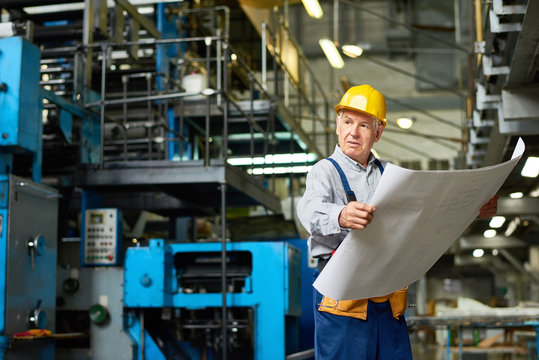 Portrait of senior man holding plans and looking away in workshop of modern factory