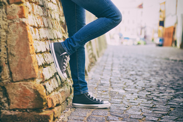 A girl wearing jeans and sneakers leans on a brick wall in the historic part of town - obrazy, fototapety, plakaty
