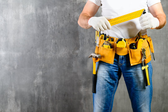 unidentified handyman standing with a tool belt with construction tools and holding roulette against grey background with copyspace. DIY tools and manual work concept