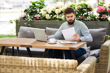 Portrait of young bearded businessman working in cafe outdoors, reading  documentation